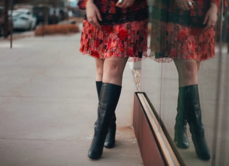 devojka u crvenoj haljini i crnim dubokim čizmama stoji naslonjena uza zid